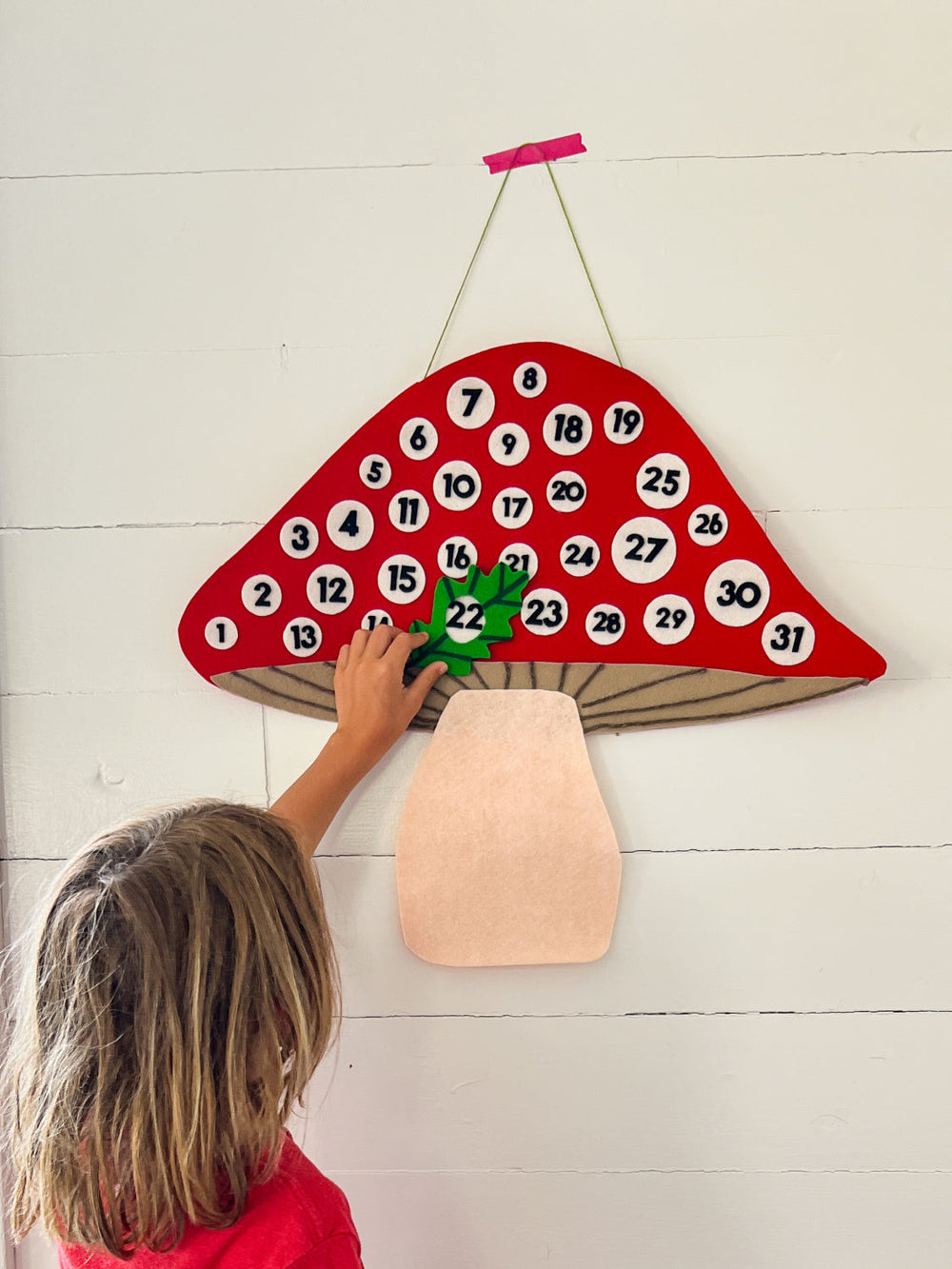Red felt mushroom calendar with white circles & black numbers. Child reaching up to put green felt around number 22.