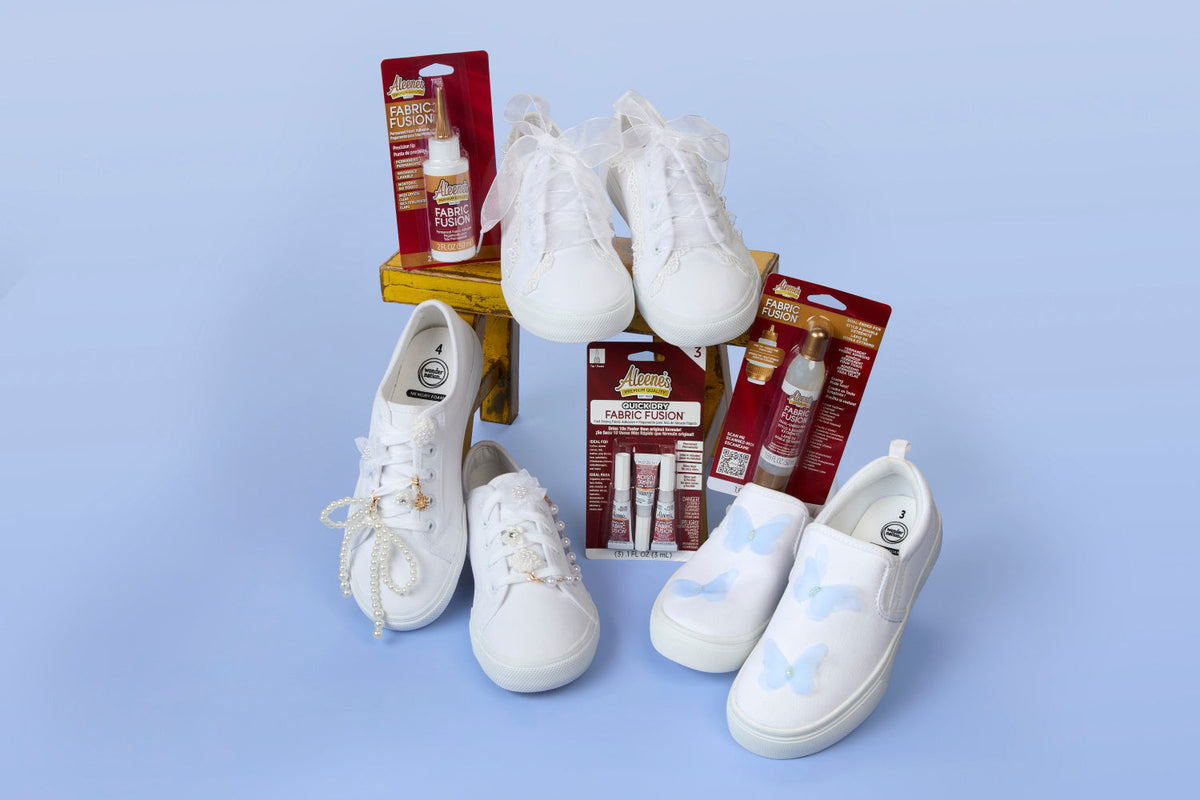 View from above of three pairs of white canvas shoes adorned with lace, pearls and fabric butterflies and surrounded by Aleene’s glues.