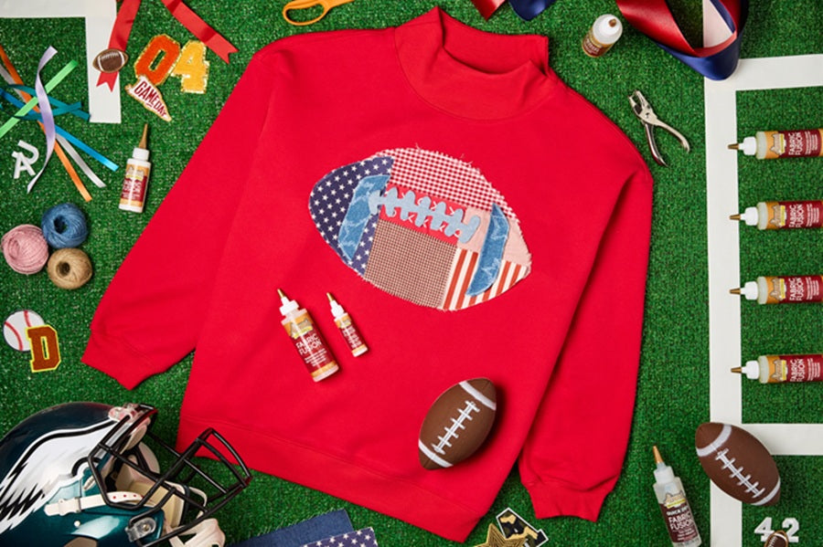 Red sweater with a patchwork football glued down in the center. Shirt is on top of a green background and surrounded by bottles of glue and football accessories.